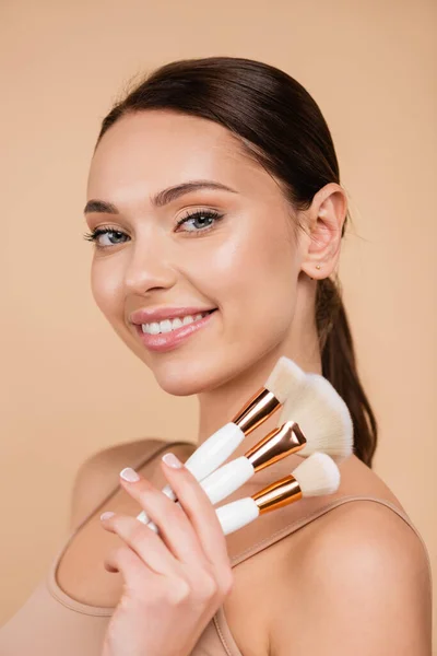 Jeune femme positive avec maquillage naturel tenant pinceaux cosmétiques isolés sur beige — Photo de stock