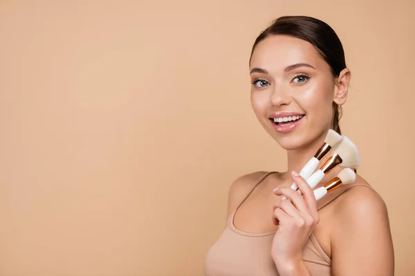 Joyeuse jeune femme avec des pinceaux cosmétiques regardant la caméra isolée sur beige — Photo de stock