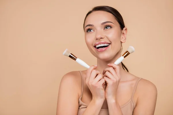 Femme excitée avec des pinceaux cosmétiques regardant loin isolé sur beige — Photo de stock