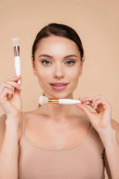 Vista frontale della giovane donna che guarda la fotocamera mentre tiene pennelli cosmetici isolati sul beige — Foto stock