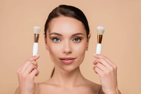 Front view of young woman with natural makeup holding cosmetic brushes isolated on beige — Stock Photo