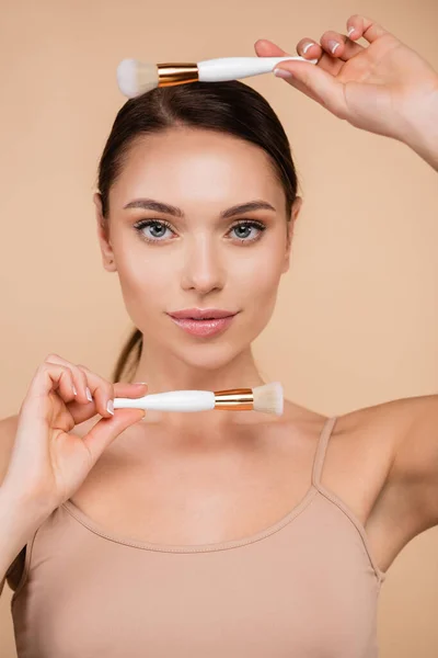 Jolie femme à la peau parfaite et au maquillage naturel posant avec des pinceaux cosmétiques isolés sur beige — Photo de stock