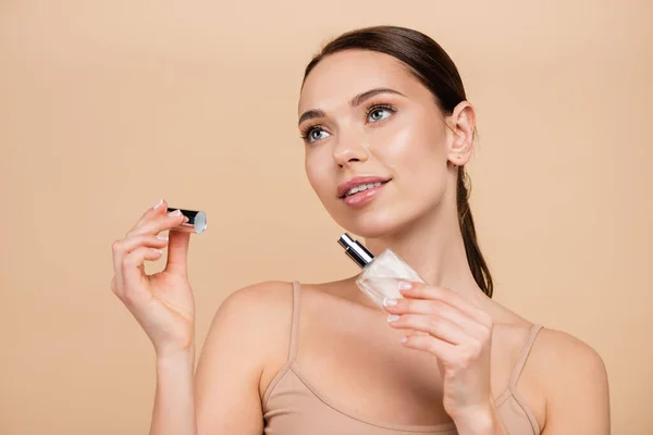 Joven mujer abriendo botella de perfume mientras mira hacia otro lado aislado en beige - foto de stock