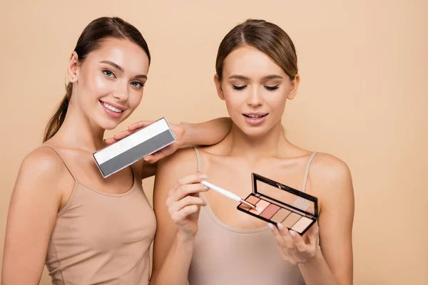 Mujeres bonitas sosteniendo paletas de luces y sombras de ojos mientras posan aisladas en beige - foto de stock