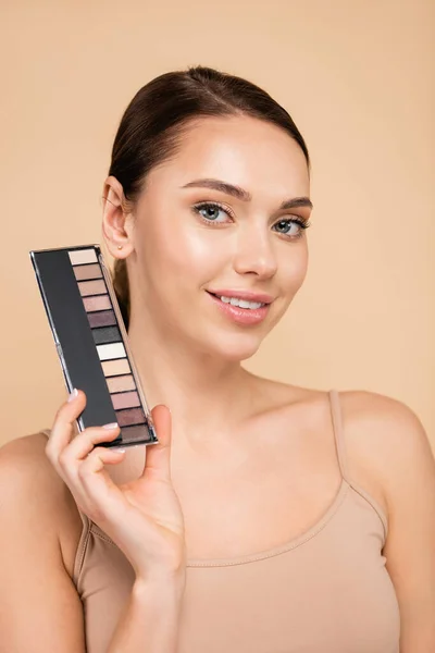 Pleased woman with eye shadows palette smiling at camera isolated on beige — Stock Photo