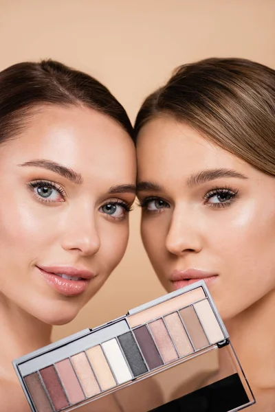 Young women with perfect skin and natural makeup looking at camera near eye shadows palette isolated on beige — Stock Photo