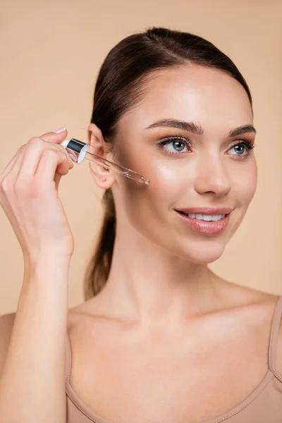 Smiling woman dropping cosmetic serum on face isolated on beige — Stock Photo