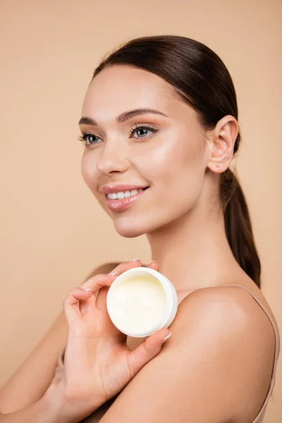 Mujer complacida sosteniendo contenedor con crema cosmética aislada en beige - foto de stock