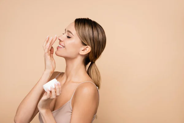 Mulher alegre aplicando creme facial no nariz isolado no bege — Fotografia de Stock