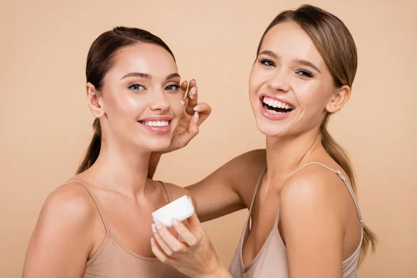 Laughing woman applying face cream on smiling friend isolated on beige — Stock Photo