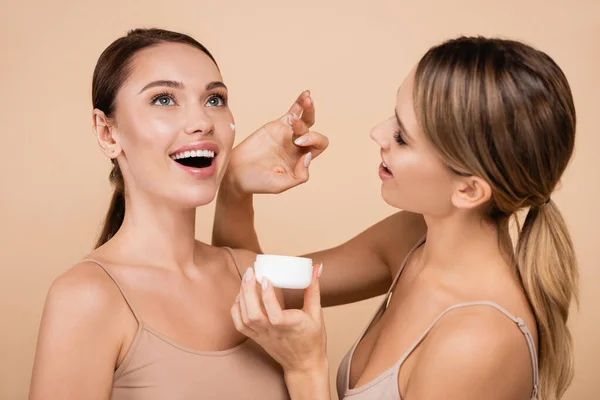 Bonita mujer aplicando crema facial en amigo excitado aislado en beige - foto de stock