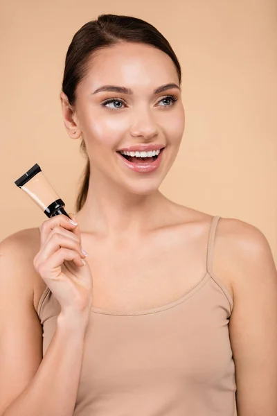 Fröhliche Frau mit Concealer-Röhre, die isoliert auf Beige wegschaut — Stockfoto