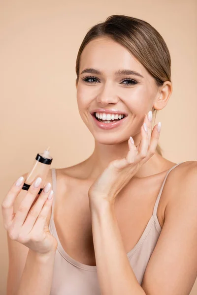 Cheerful woman applying face foundation while looking at camera isolated on beige — Stock Photo