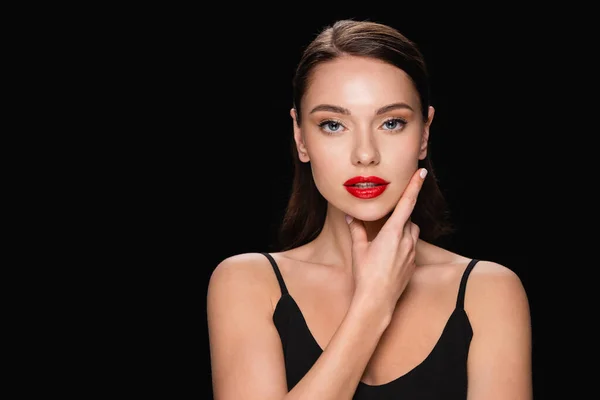 Sinnliche Frau mit roten Lippen, die Gesicht berühren, während sie die Kamera isoliert auf schwarz betrachtet — Stockfoto