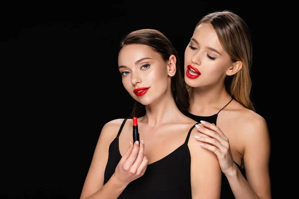 Sensual woman touching shoulder of pretty friend holding red lipstick isolated on black — Stock Photo