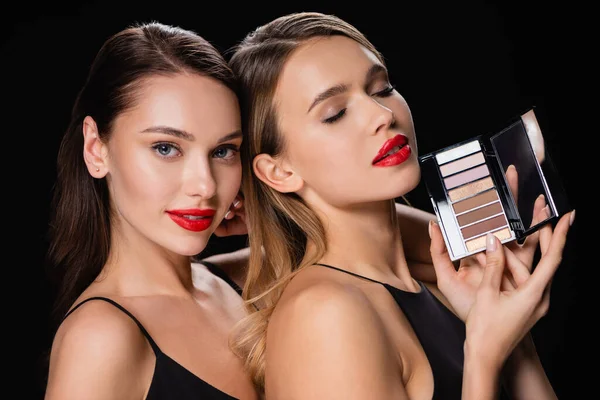 Sensual woman looking at camera near pretty friend with eye shadows palette isolated on black — Stock Photo