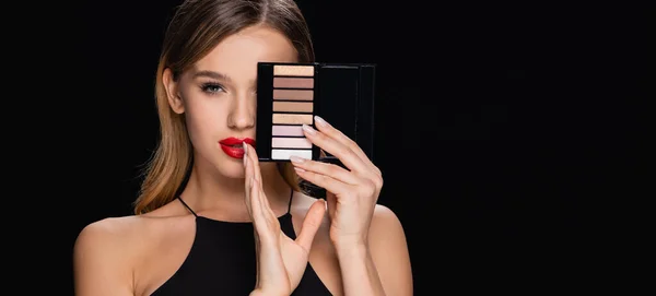 Young woman with red lips obscuring face with eye shadows palette isolated on black, banner — Stock Photo