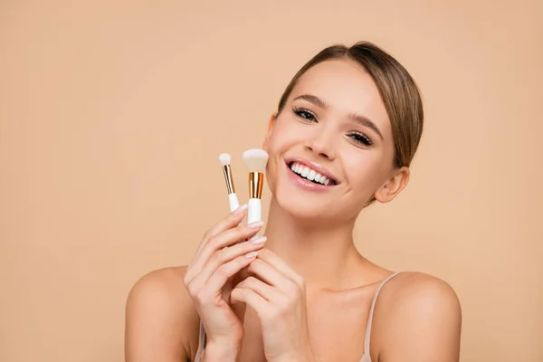 Fröhliche Frau mit Kosmetikpinseln, die isoliert auf Beige in die Kamera lächelt — Stockfoto