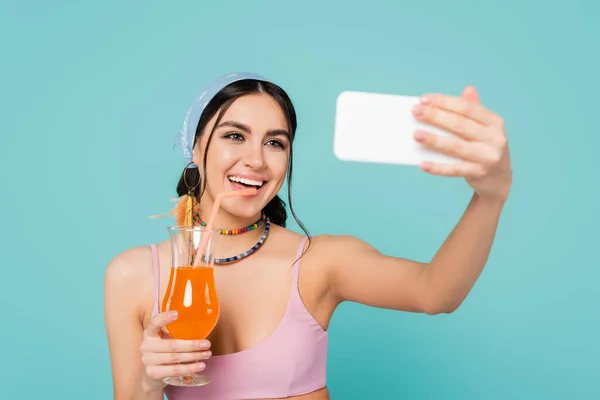 Positive woman with cocktail taking selfie on blurred smartphone isolated on blue — Stock Photo