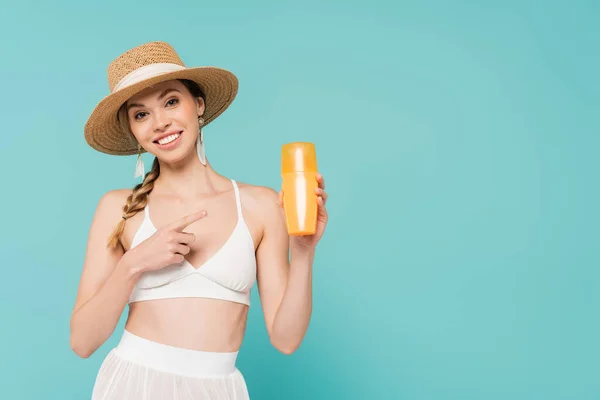 Позитивна жінка в сонячному капелюсі вказує на сонцезахисний крем ізольовано на синьому — стокове фото