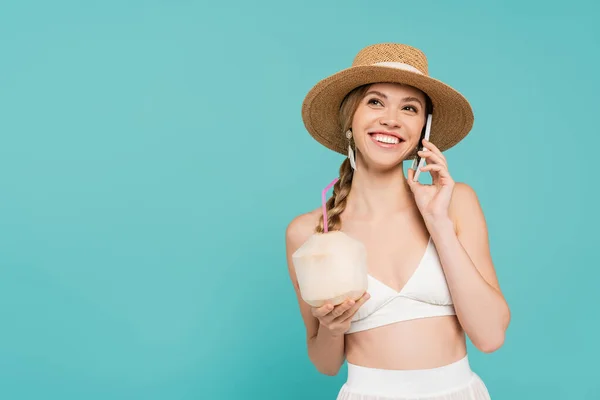 Mujer alegre en sombrero de sol hablando en el teléfono inteligente y la celebración de cóctel en coco aislado en azul - foto de stock