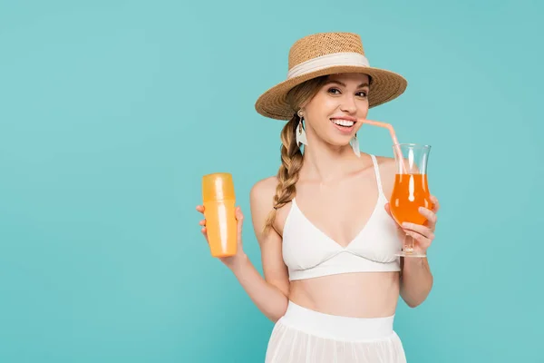 Femme souriante en chapeau de paille tenant crème solaire et cocktail isolé sur bleu — Photo de stock