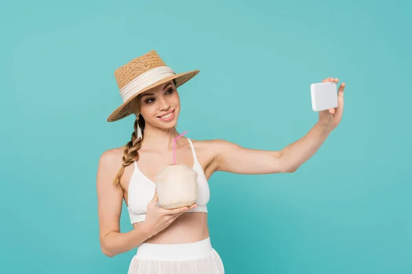 Mujer sonriente en sombrero de sol celebración de cóctel en coco y tomar selfie en el teléfono inteligente aislado en azul - foto de stock