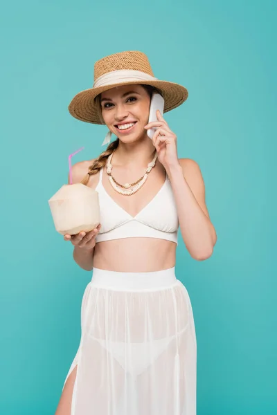Позитивна жінка в сонячному капелюсі говорить на смартфоні і тримає коктейль в кокосі ізольовано на синьому — стокове фото