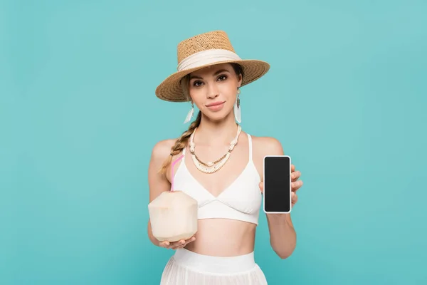 Молодая женщина в шляпе и топ держа смартфон и коктейль в кокосе изолированы на синий — стоковое фото