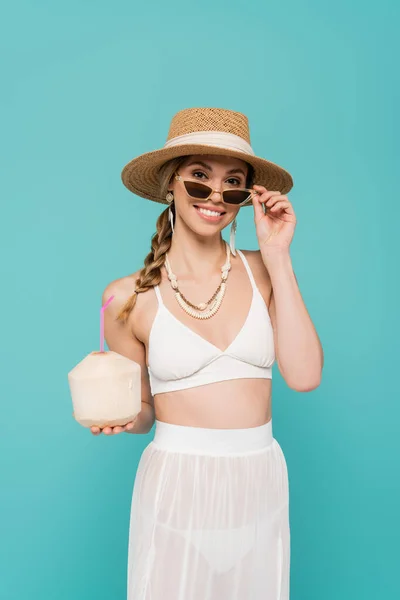 Mujer positiva en sombrero de sol y traje de baño celebración de cóctel en coco aislado en azul - foto de stock