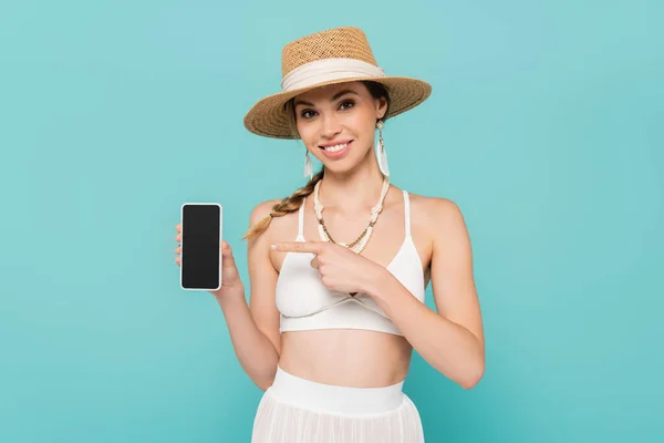 Усміхнена жінка в сонячному капелюсі вказує на смартфон ізольовано на синьому — стокове фото