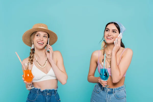 Mulheres felizes em tops segurando coquetéis e falando em smartphones isolados em azul — Fotografia de Stock