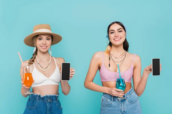 Mulheres sorridentes em tops e colares segurando smartphones e coquetéis isolados em azul — Fotografia de Stock