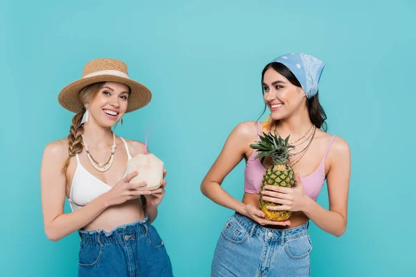 Mulheres sorridentes em tops segurando coquetel em coco e abacaxi isolado em azul — Fotografia de Stock