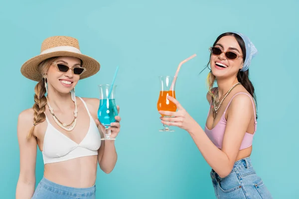 Mujeres positivas en tops y gafas de sol sosteniendo cócteles aislados en azul - foto de stock