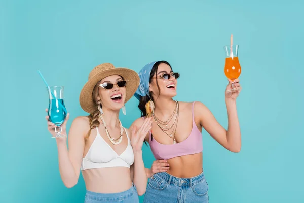 Mujeres alegres en gafas de sol sosteniendo cócteles aislados en azul - foto de stock