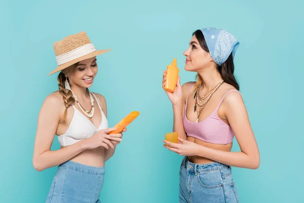 Mujer sonriente en sombrero de sol sosteniendo protector solar cerca de amigo aislado en azul - foto de stock