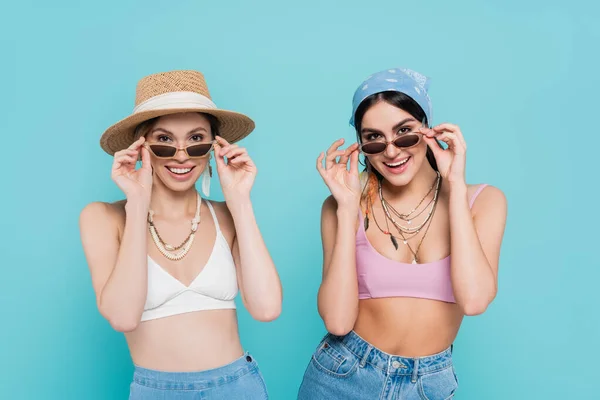 Positive women in tops holding sunglasses isolated on blue — Stock Photo
