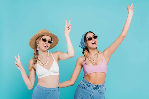 Mulheres felizes em topos e óculos de sol mostrando gesto de paz isolado em azul — Fotografia de Stock