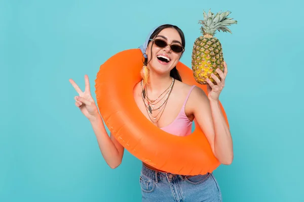 Donna positiva in anello di nuoto tenendo ananas e mostrando segno di pace isolato su blu — Foto stock