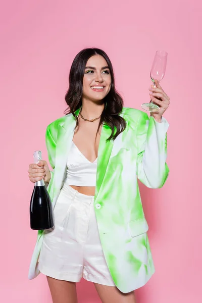 Smiling woman holding glass and bottle of champagne on pink background — Stock Photo