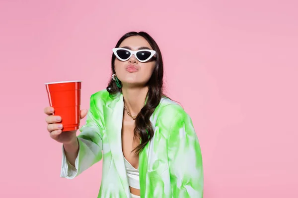 Femme élégante dans des lunettes de soleil tenant tasse en plastique et lèvres boudantes isolées sur rose — Photo de stock