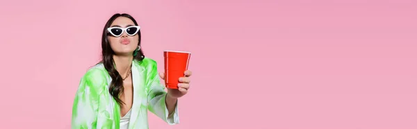 Mujer joven en gafas de sol haciendo pucheros labios y sosteniendo taza de plástico aislado en rosa, pancarta - foto de stock