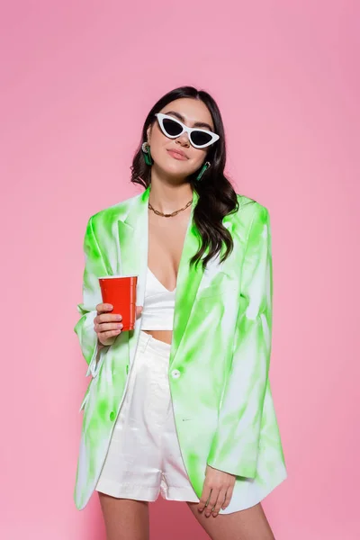 Jeune femme en blazer et lunettes de soleil tenant tasse en plastique sur fond rose — Photo de stock