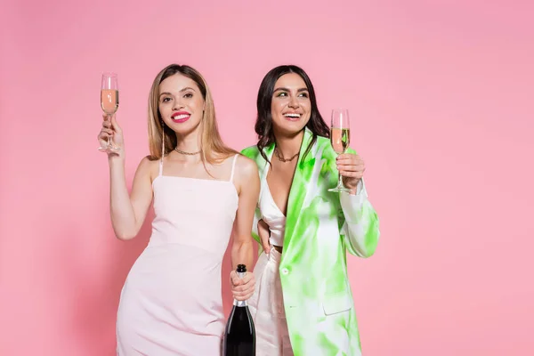 Positive women holding glasses and bottle of champagne on pink background — Stock Photo