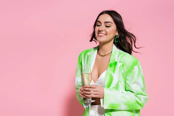 Donna positiva in collana e giacca in possesso di un bicchiere di champagne su sfondo rosa — Foto stock