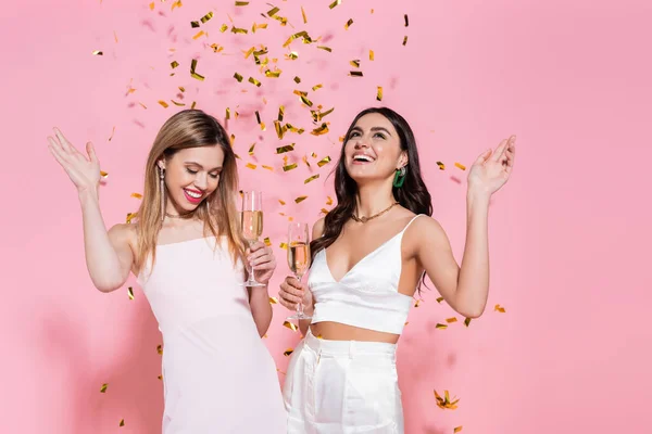 Pretty friends holding champagne under falling confetti on pink background — Stock Photo