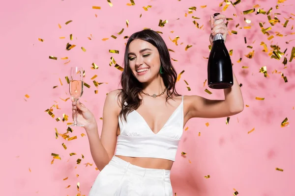 Красивая брюнетка держит бутылку и бокал шампанского под золотыми конфетти на розовом фоне — стоковое фото