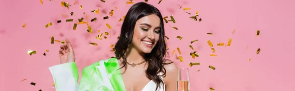 Pretty woman smiling while looking at glass of champagne near confetti on pink background, banner — Stock Photo