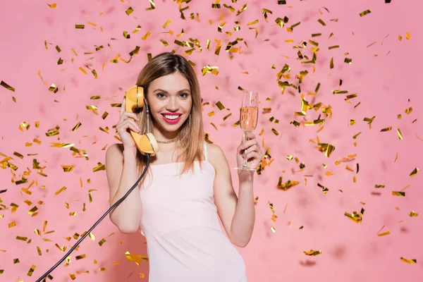 Cheerful woman with champagne talking on telephone under confetti on pink background — Stock Photo